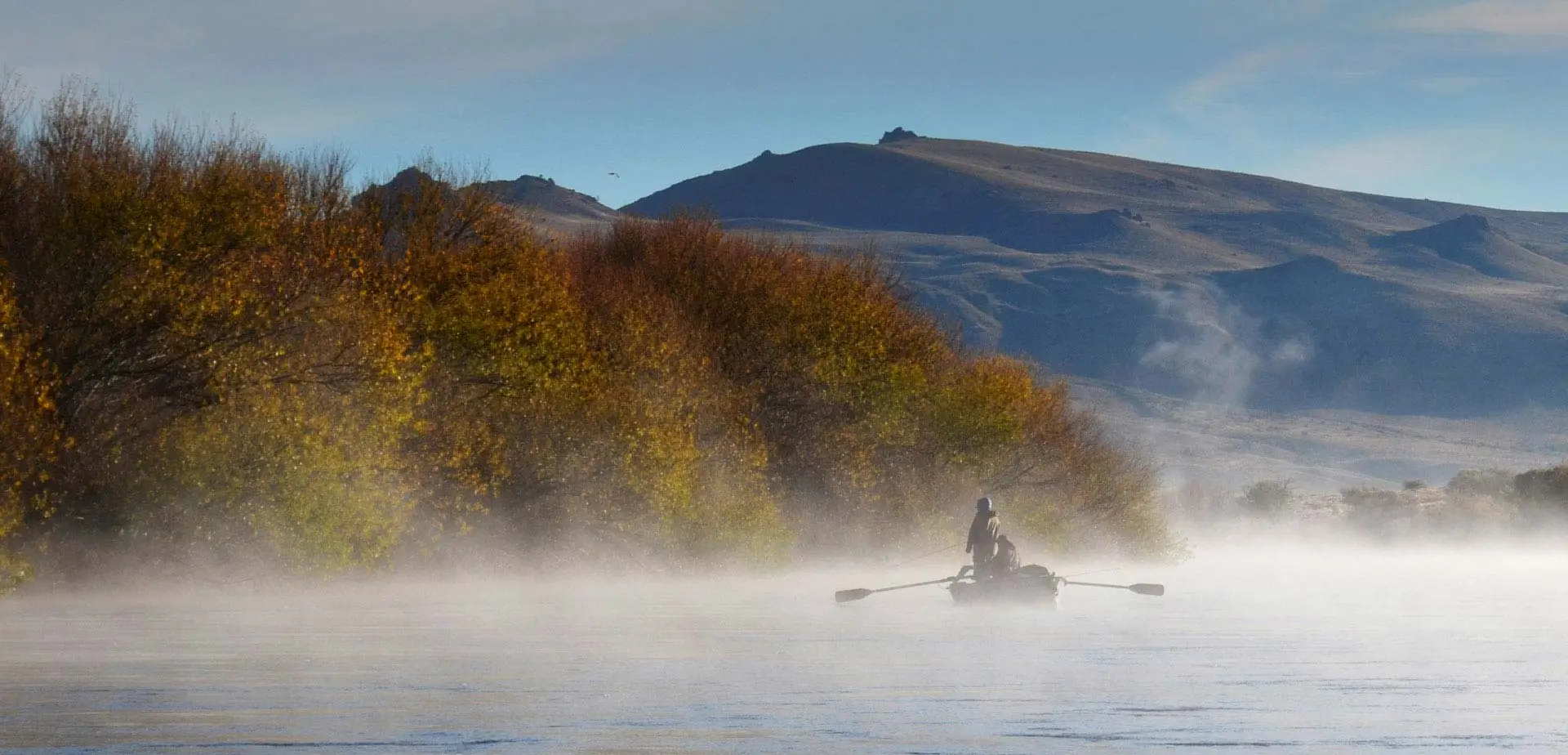 Barco a la deriva navegando por el brumoso río Limay al amanecer durante un tour diario de pesca con mosca en Bariloche, Patagonia Argentina, rodeado de árboles y montañas de colores otoñales.