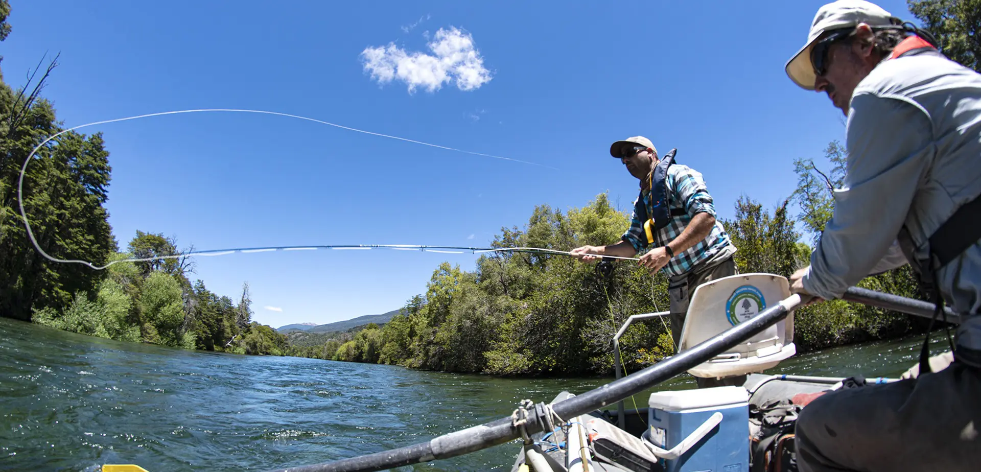 Guía de pesca con mosca dirigiendo el barco mientras el cliente lanza una línea en un río Manso durante un viaje guiado en flotador en Bariloche, Patagonia Argentina
